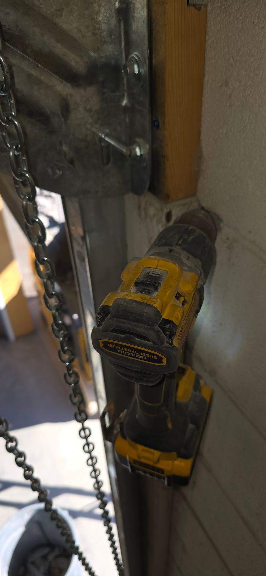 Yellow cordless drill hanging on a chain beside a wall and wooden beam.