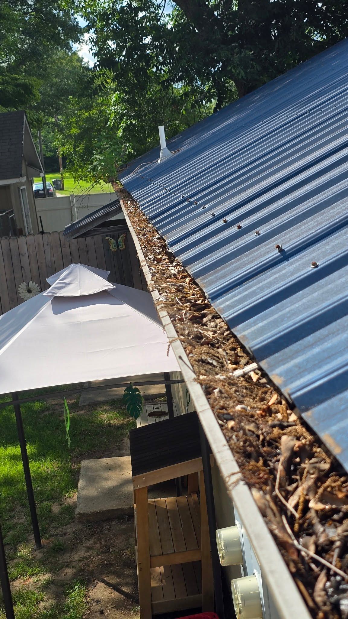 Blue metal roof with a gutter full of leaves beside a patio umbrella and trees