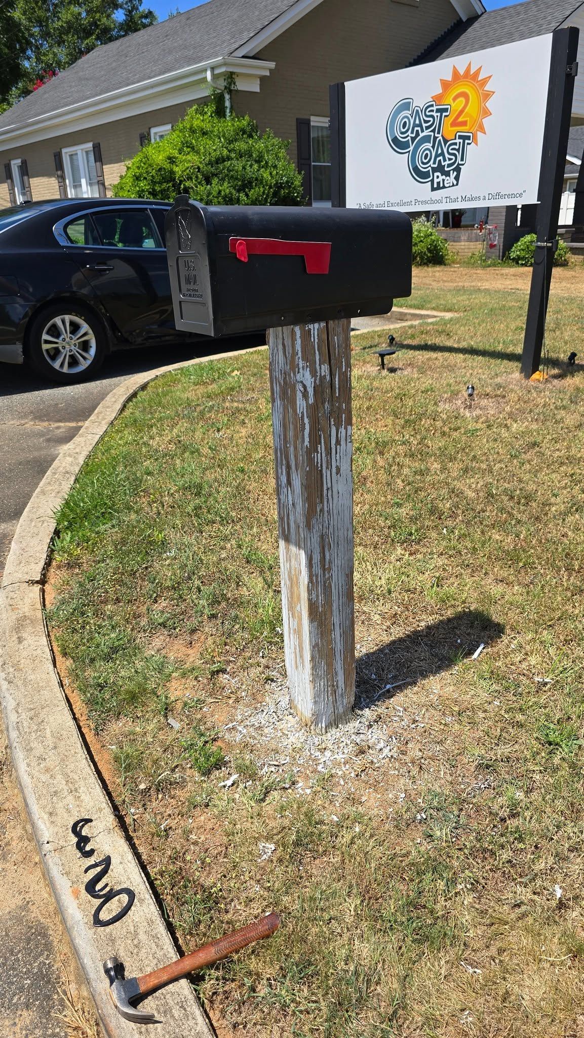 Black mailbox on a post by a driveway with a parked car and house sign in the background
