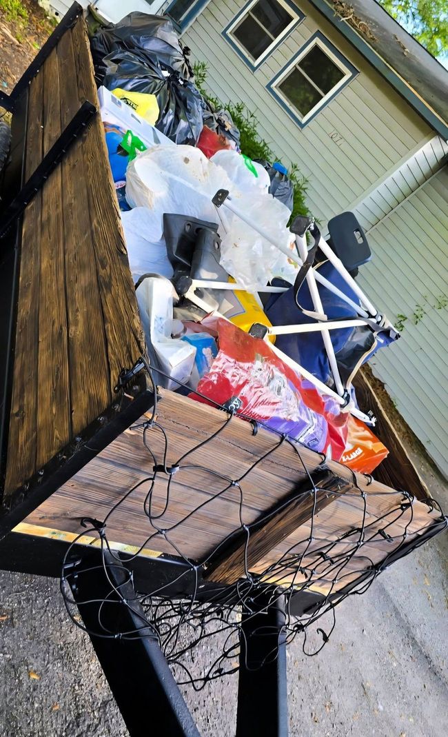 Overflowing black trash bin with bags, cardboard, and colorful debris on a wet driveway beside a house
