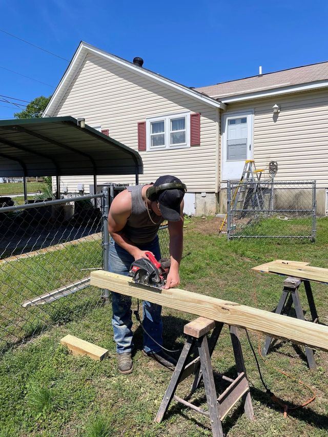 Person using a circular saw to cut a long board on sawhorses in a backyard beside a house.