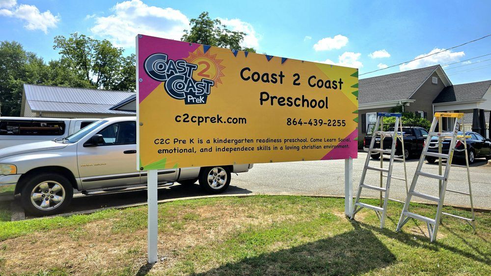 Coast 2 Coast Preschool sign on a pink-yellow banner in a grassy yard with vehicles and tents nearby