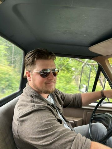 Man in sunglasses sitting in the driver’s seat of an older vehicle, looking at the camera.
