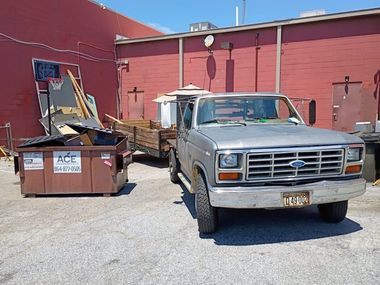 Old silver pickup truck beside a dumpster and trailer in a red-walled alley