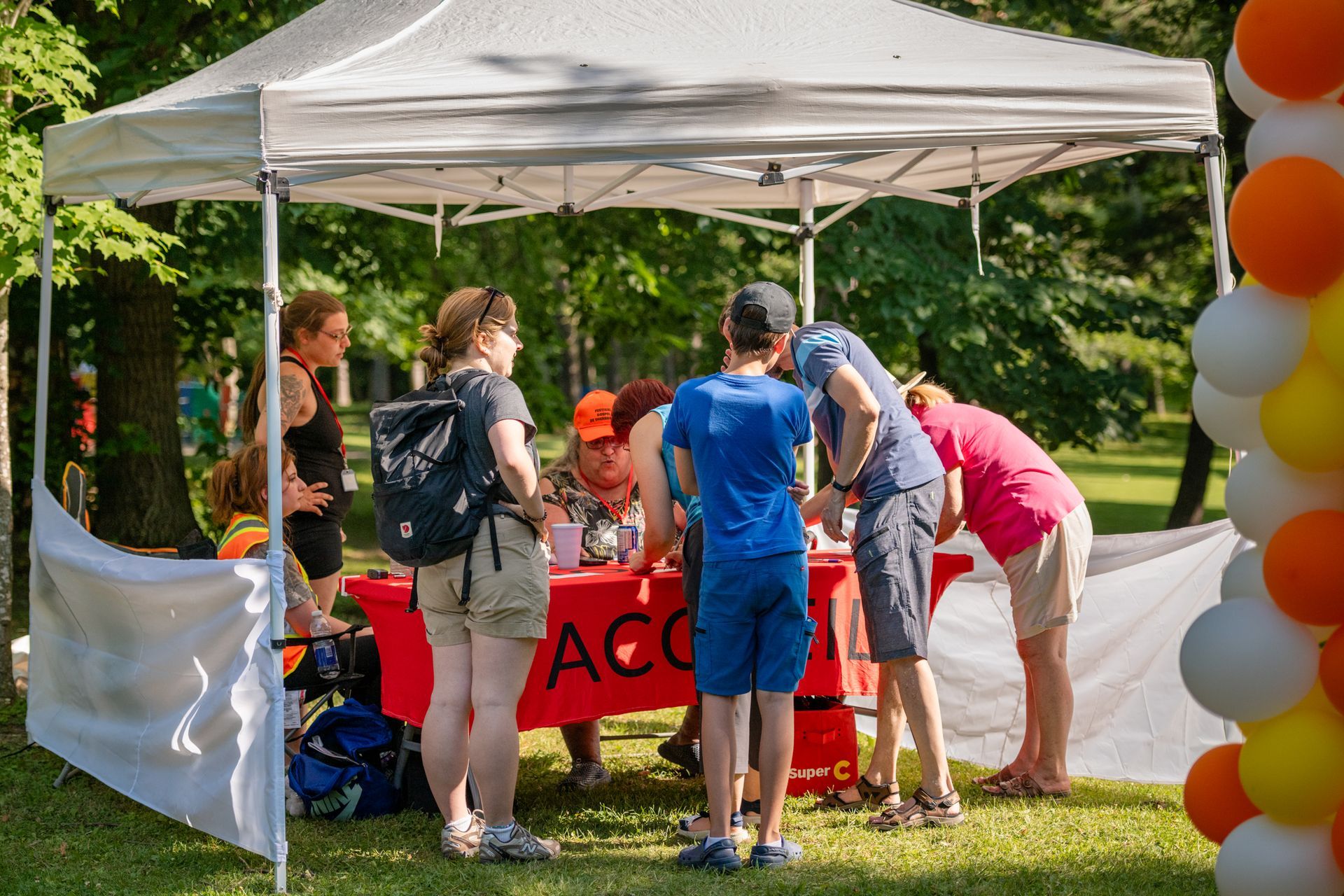 Des personnes étaient rassemblées sous une tente blanche avec une table rouge lors d'un événement en plein air, près d'une colonne de ballons.