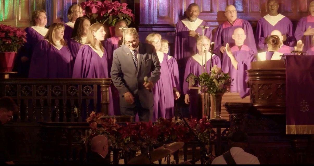 À VENIR Chœur et orateurs en robes violettes sur la scène d'une église, avec un homme et une femme debout au centre sous une lumière chaude