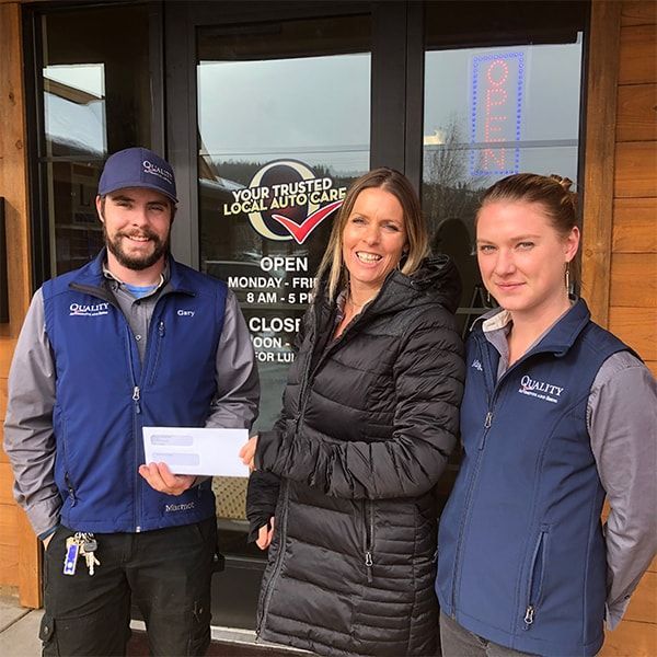 Three people standing outside a storefront, smiling and holding a large check | Quality Automotive Servicing