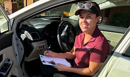 Person smiling in a car doorway, holding papers, with the driver's seat and dashboard visible | Quality Automotive Servicing