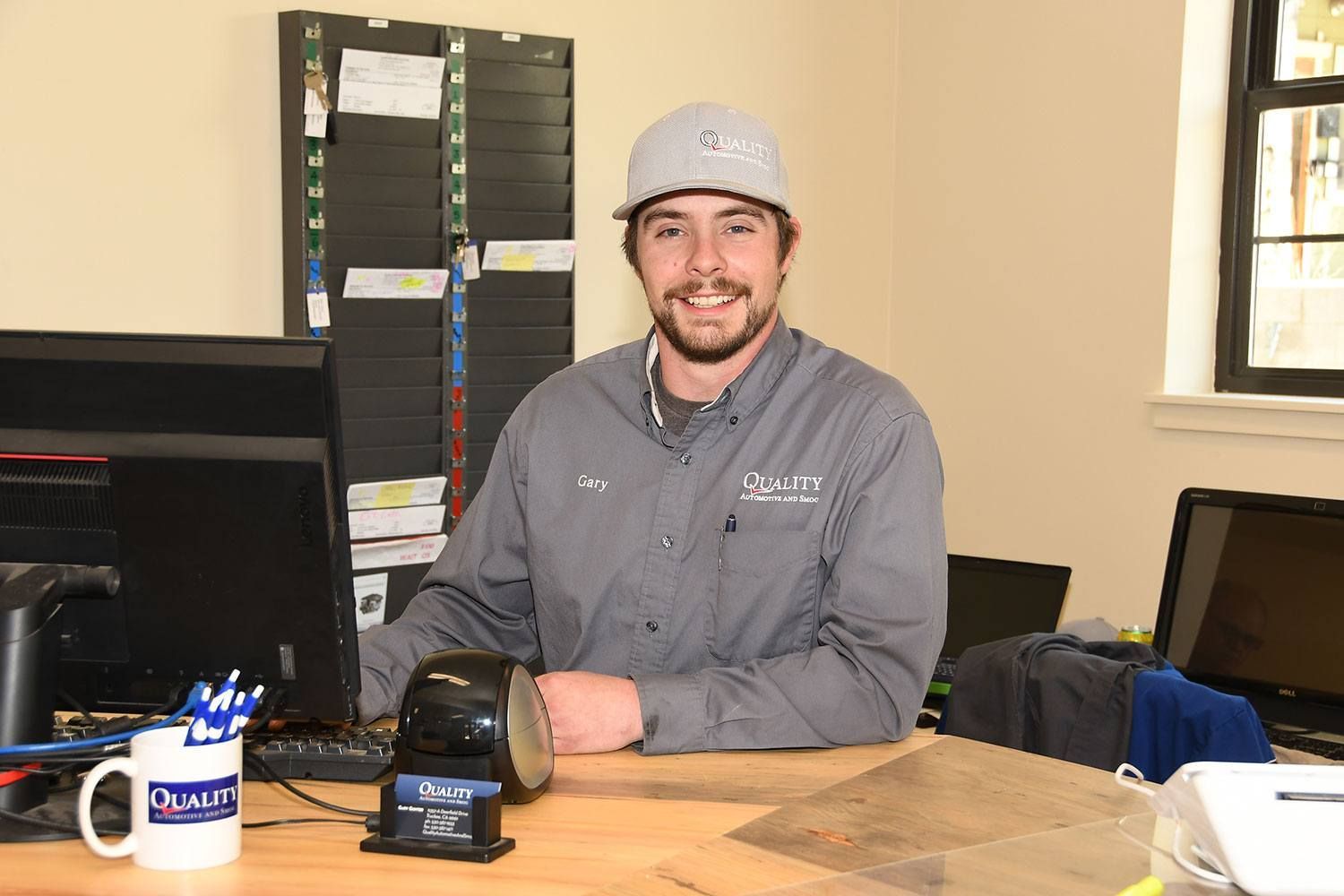 Man in a gray shirt and cap sitting at a desk with a laptop in an office, smiling | Quality Automotive Servicing