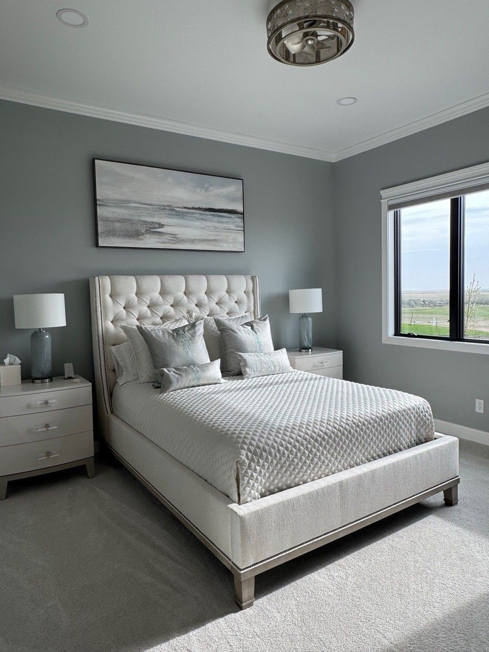 Bright gray bedroom with tufted bed, nightstands, and large window overlooking a landscape