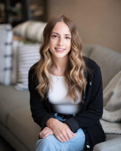 Smiling woman seated on a couch, wearing a black cardigan, white top, and light jeans.