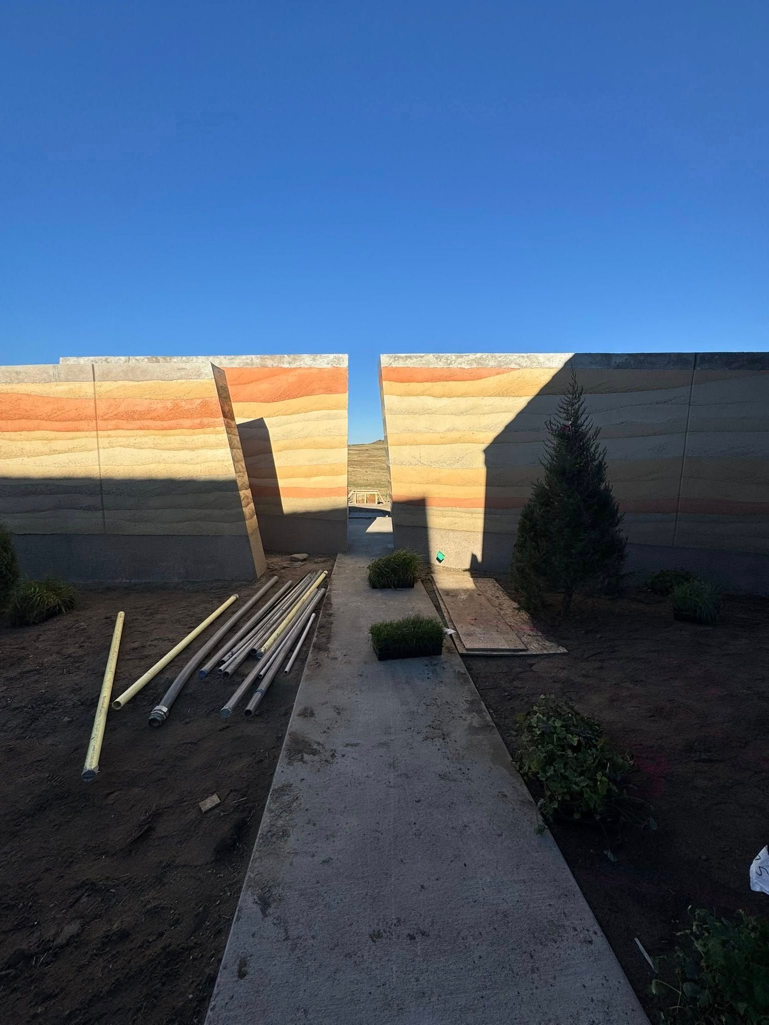 Backyard walkway between tall brick walls under a clear blue sky, with potted plants and pipes on the left.