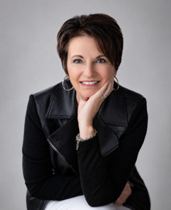 Smiling person with short dark hair in a black jacket, resting chin on hand against a gray background