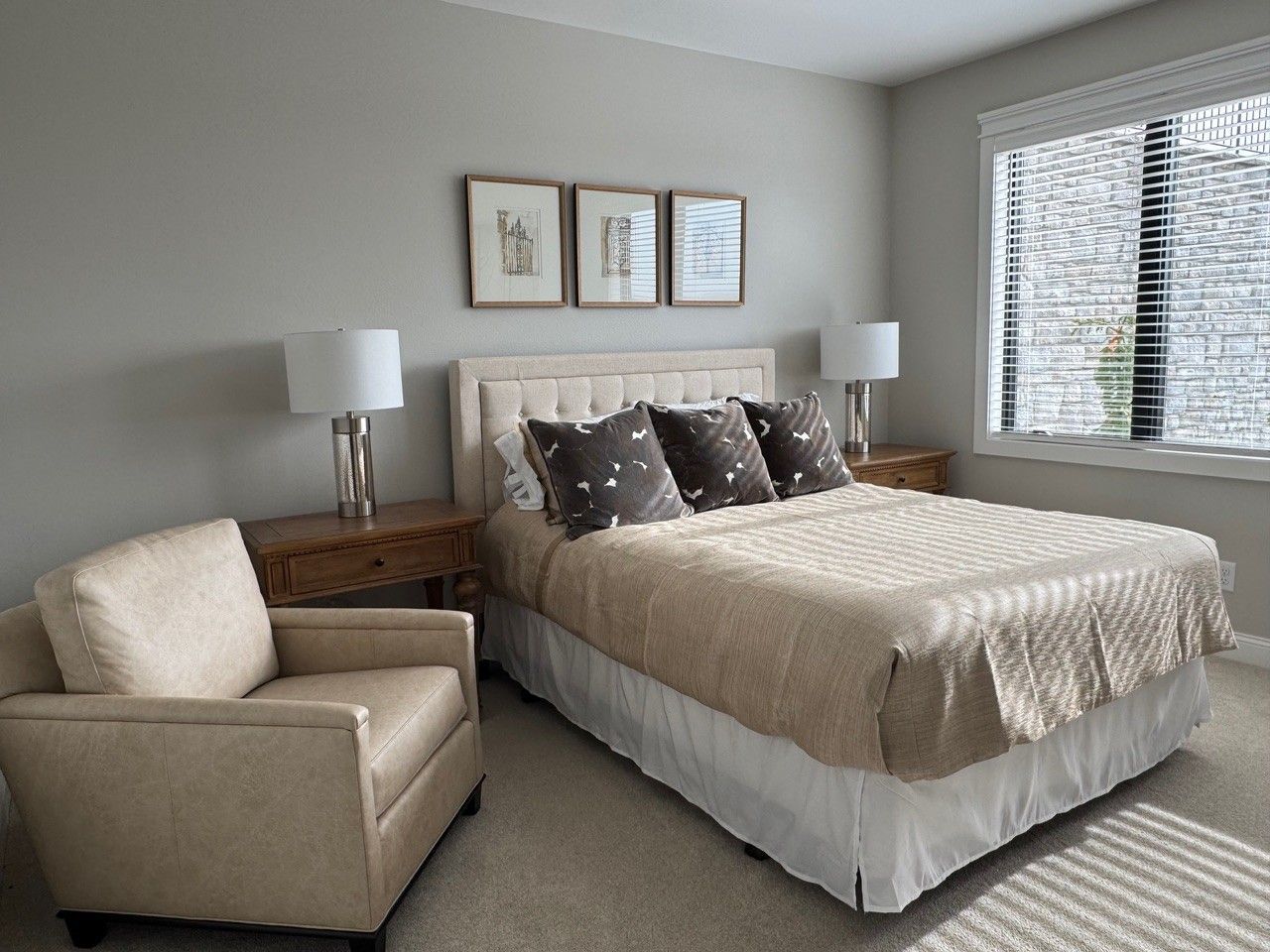 Cozy bedroom with beige bed, two lamps, armchair, and framed art on a light gray wall.