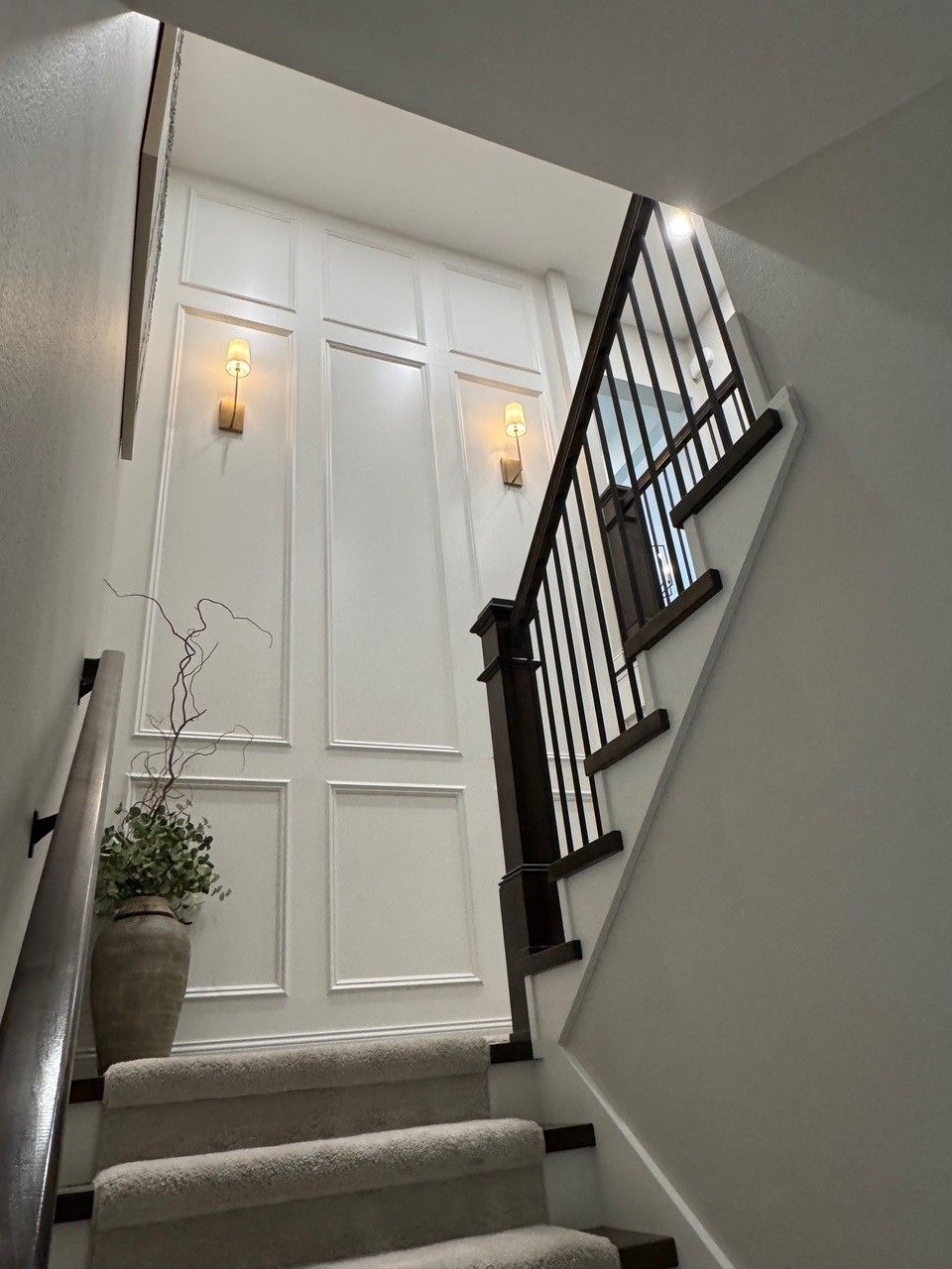 Narrow staircase with gray carpet, black railing, white paneled wall, and warm wall sconces.