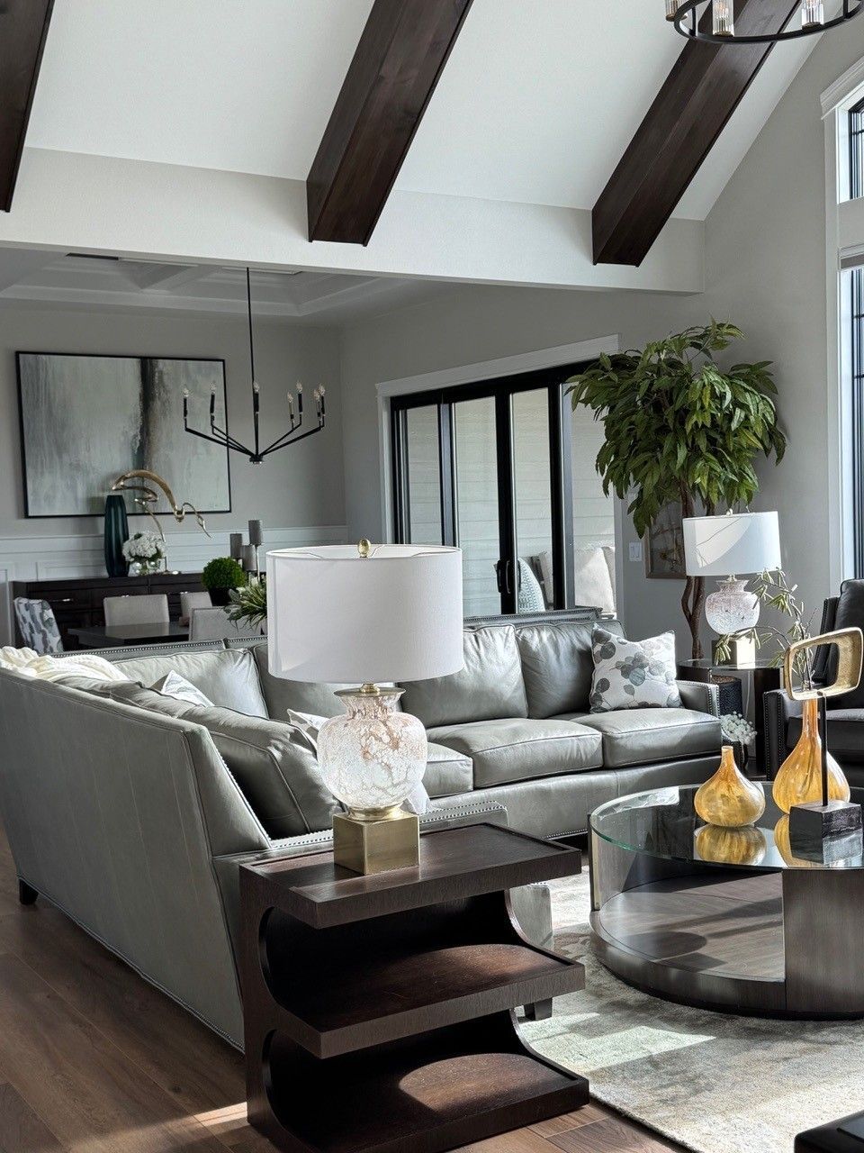Modern living room with gray sectional sofa, wood beams, large windows, and round coffee table.