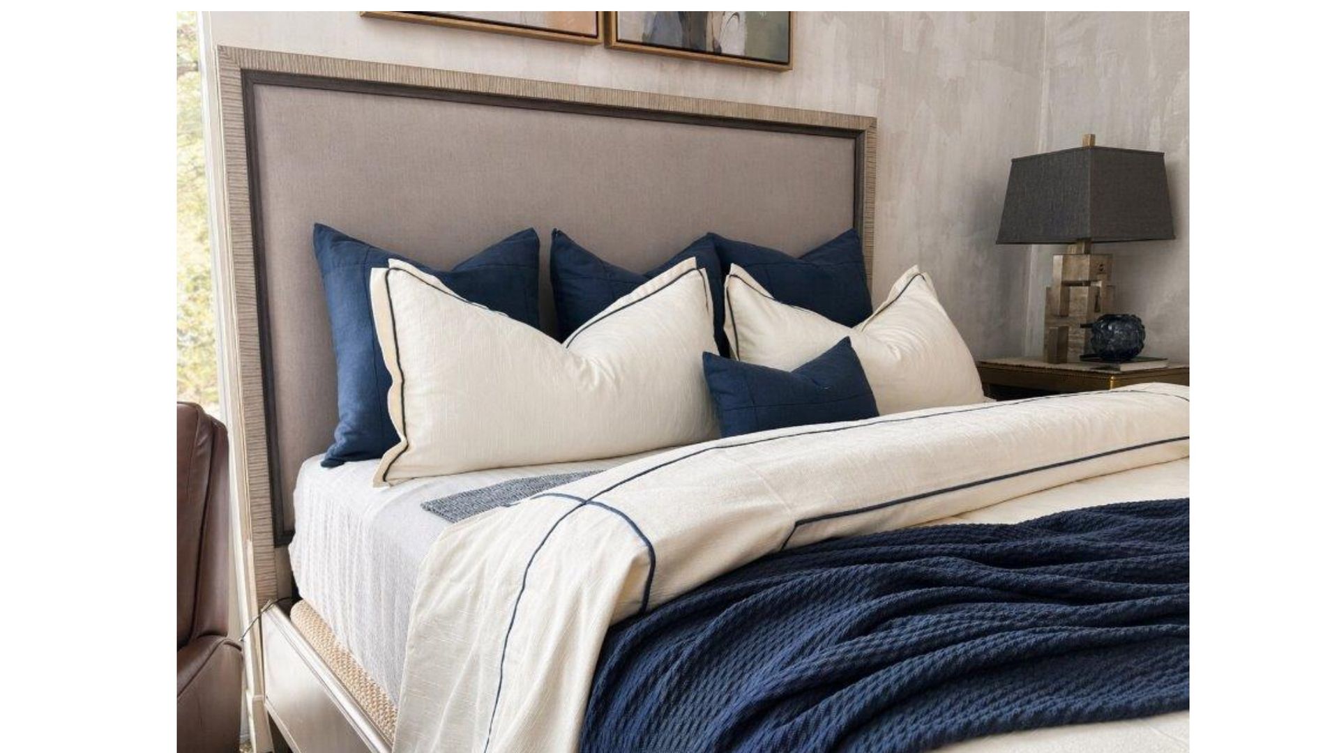 Neatly made bed with beige headboard, white bedding, and navy accent pillows in a bright bedroom