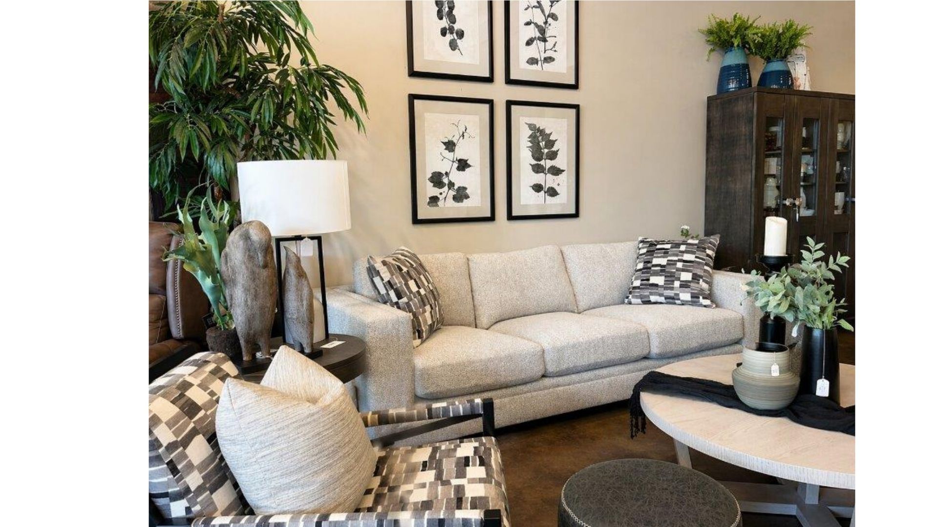 Modern living room with beige sofa, patterned chairs, wall art, plants, and a dark cabinet.