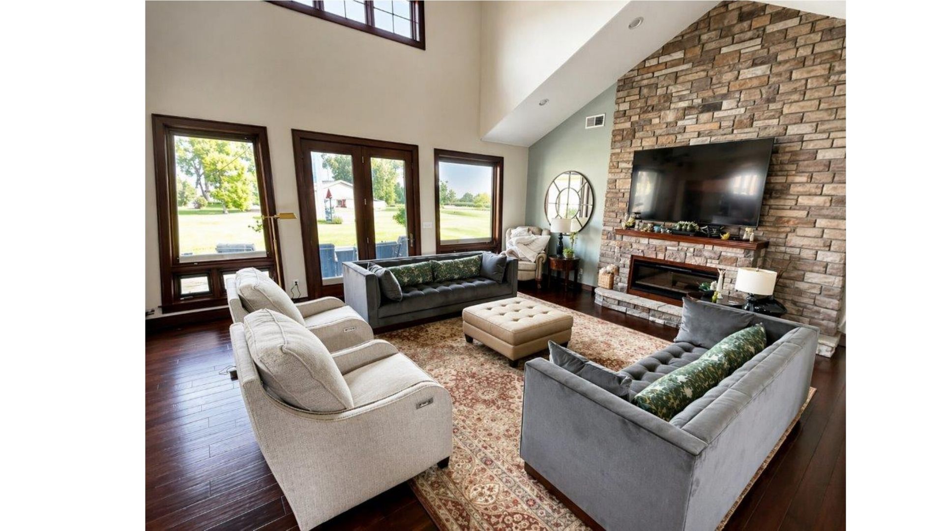 Modern living room with stone fireplace, wall-mounted TV, and gray and white sofas around a tufted ottoman.