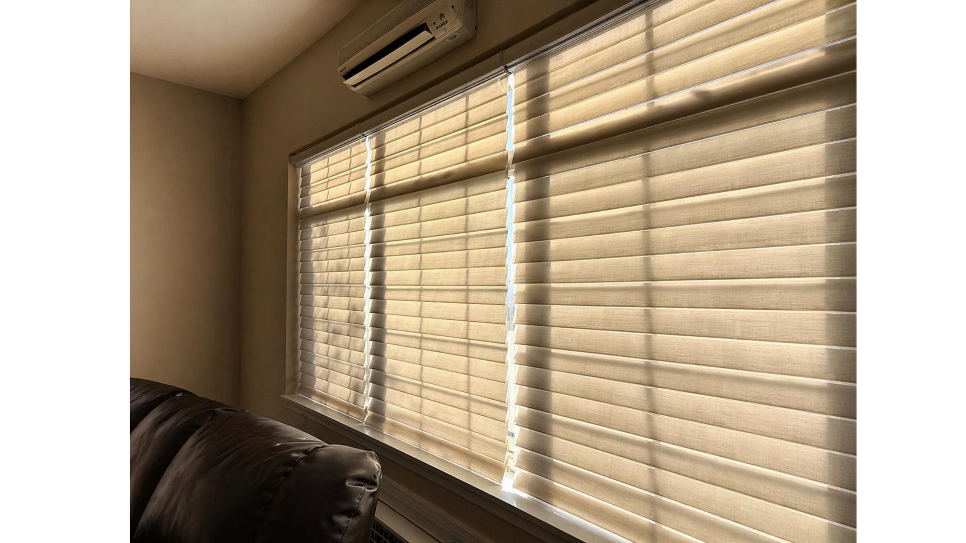 Sunlit living room with closed beige blinds and a wall-mounted air conditioner above a dark sofa