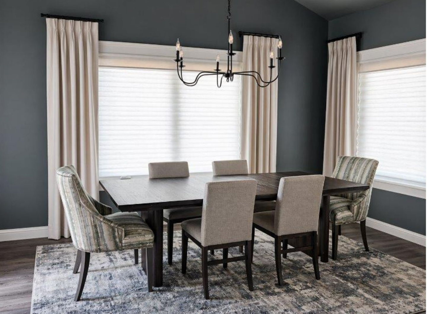 Dining room with dark table, six upholstered chairs, chandelier, and large windows with blinds and curtains