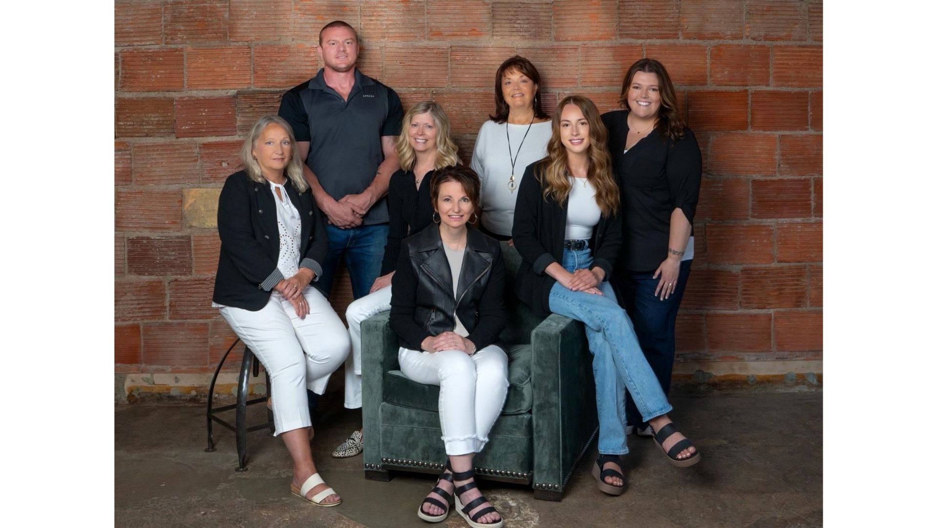 Six people posed for a group portrait indoors against a brick wall, seated and standing on white chairs.