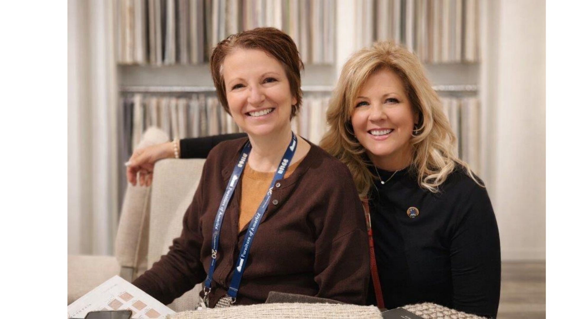 Two smiling women sitting on a couch indoors, wearing lanyards and dark clothing.