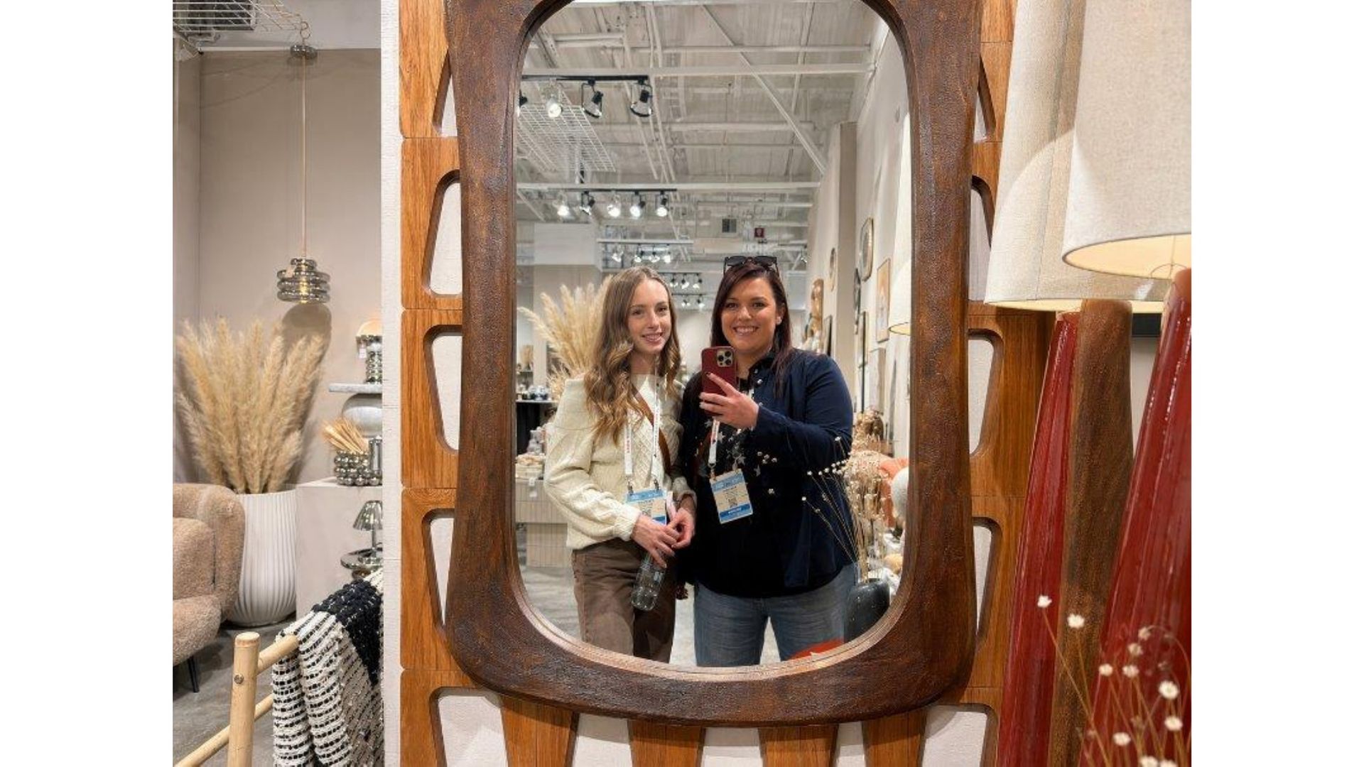Two people posing in a mirror inside a home decor store, with woven and wooden furnishings around them.