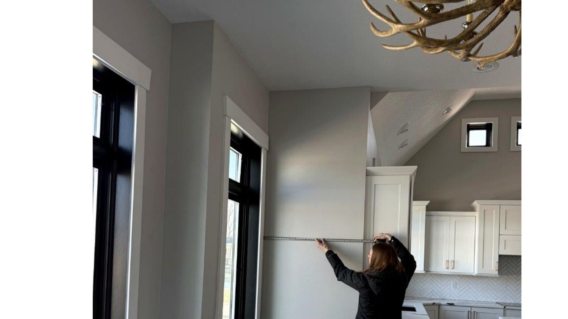 Person measuring a wall in a bright gray room with windows and built-in cabinets