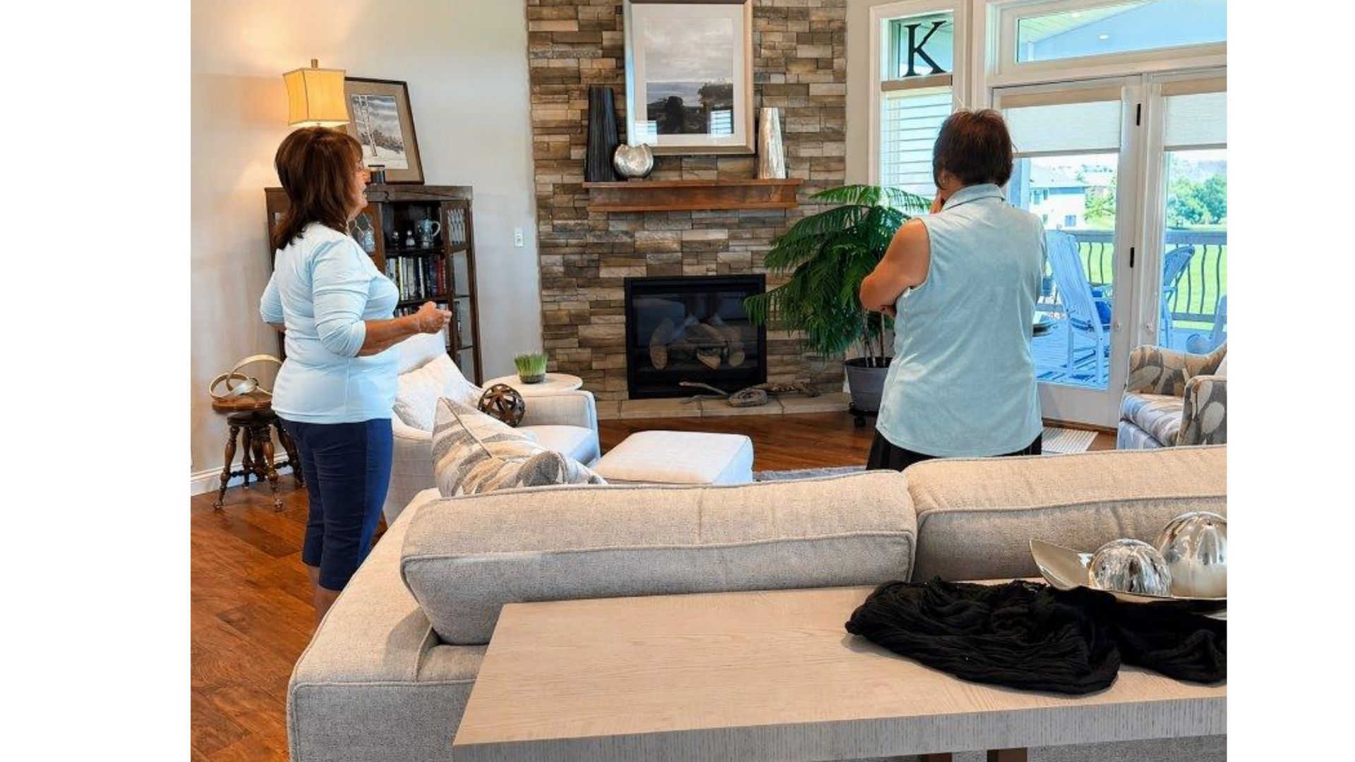 Two people in a bright living room, arranging decor near a stone fireplace and large windows.