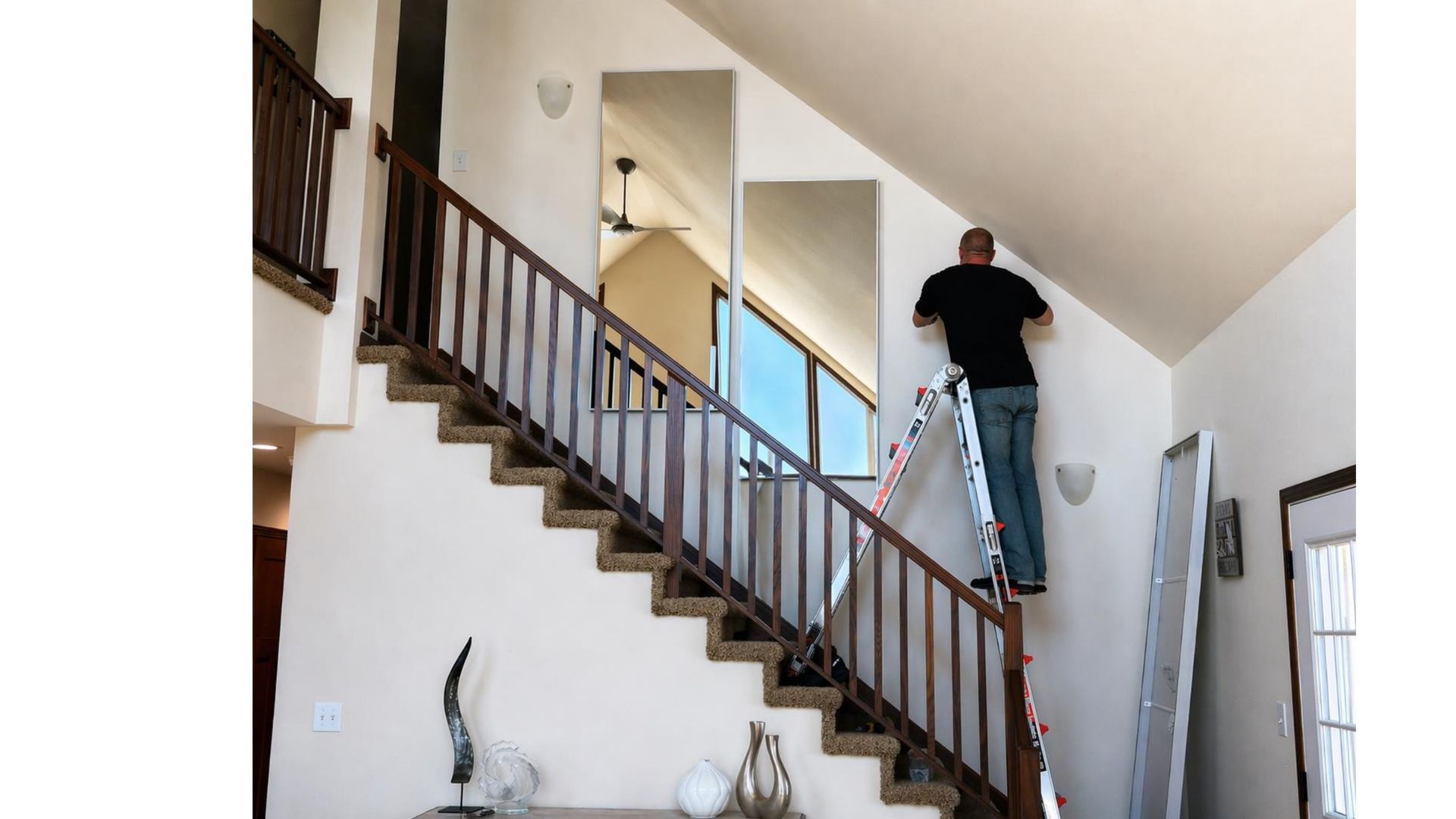 Man standing on a ladder painting a white stairwell with dark wood railings and a sloped ceiling