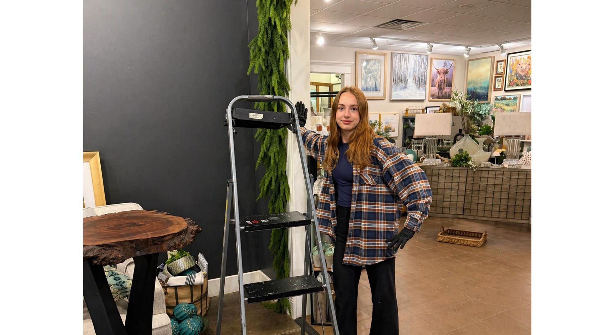 Woman standing beside a ladder in a plant-filled shop or greenhouse with framed art on the walls