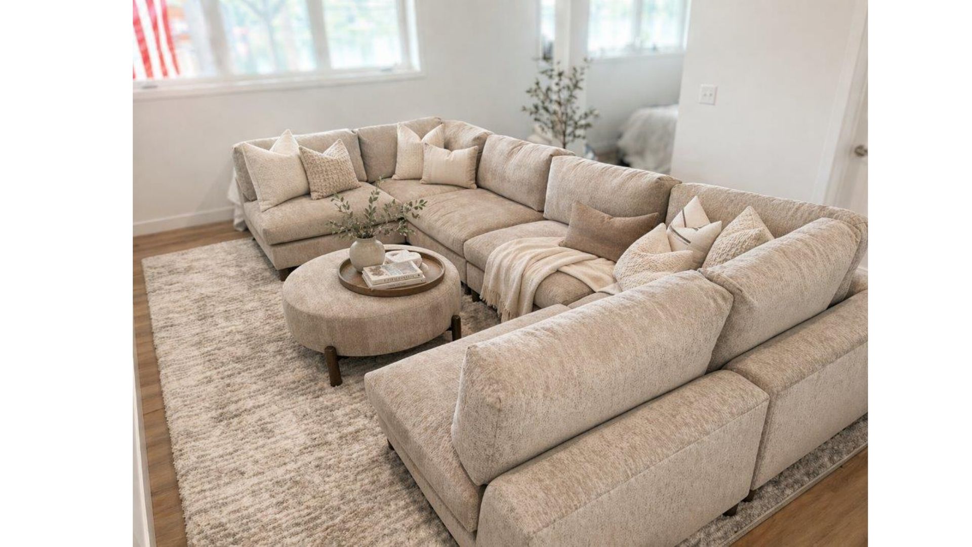 Beige sectional sofa with pillows around a round ottoman in a bright, carpeted living room