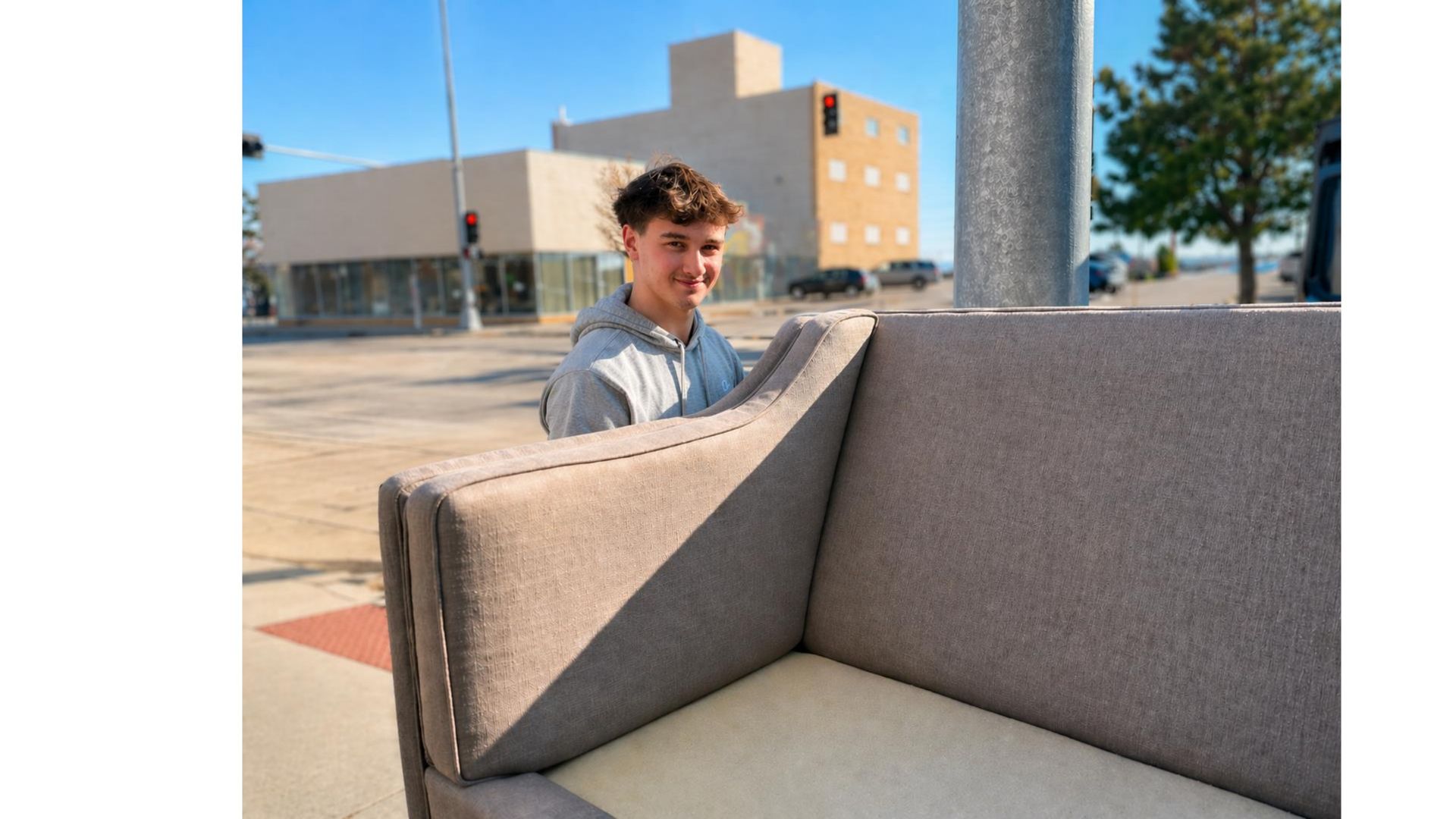 Young person standing behind a gray outdoor couch in a sunny plaza near a building.