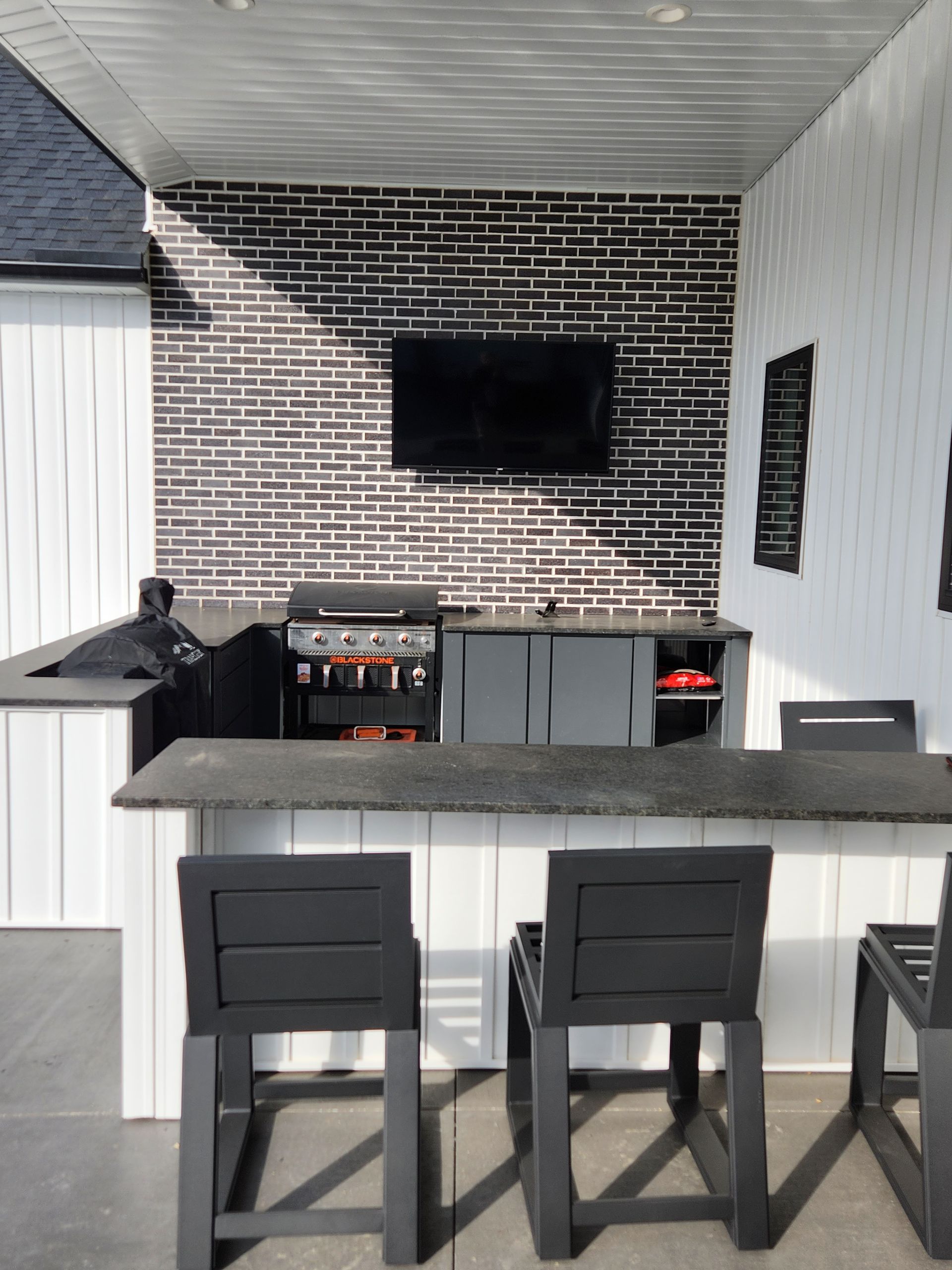 Outdoor kitchen with a black brick accent wall, TV, and gray counter with bar stools.