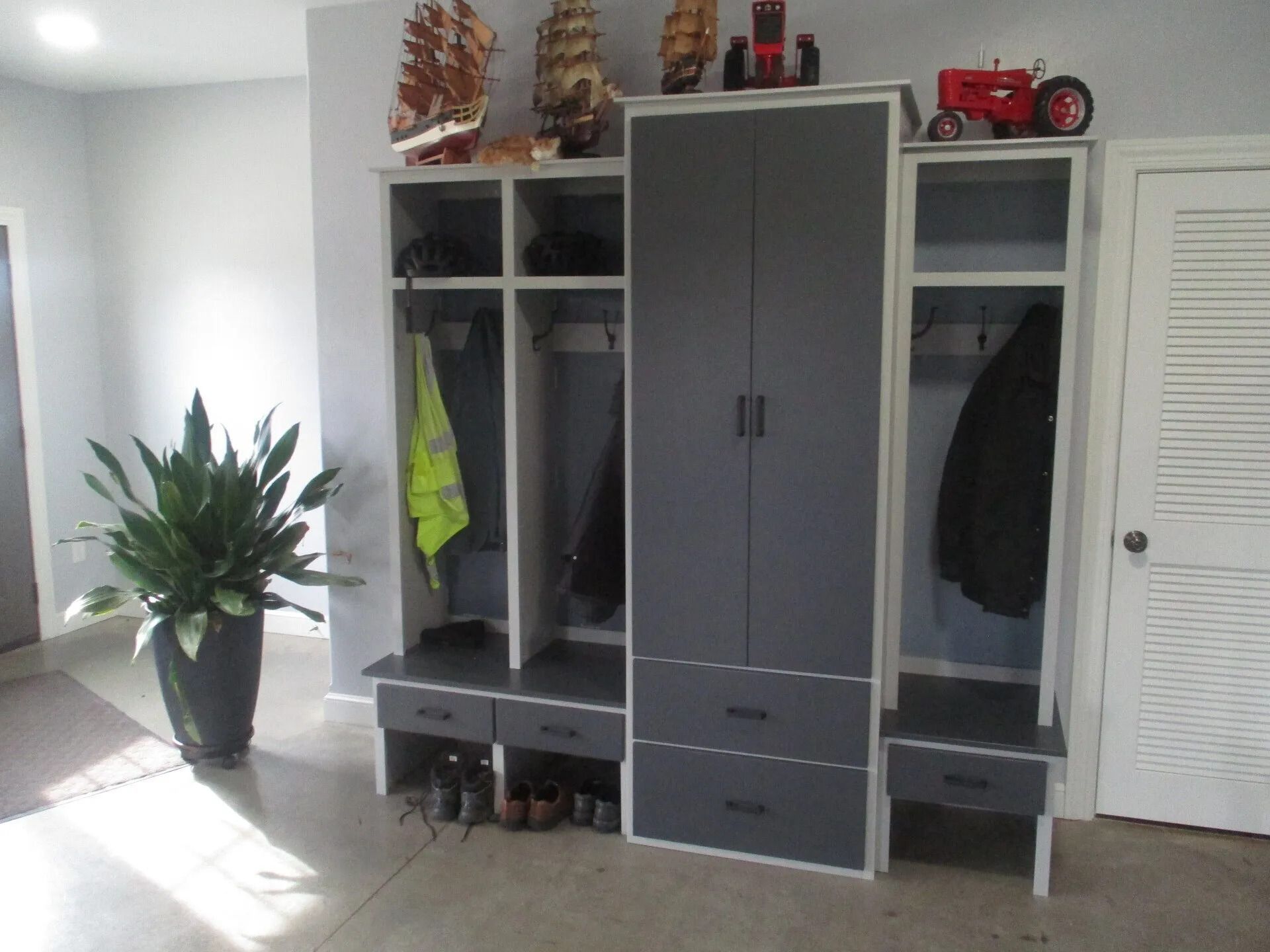 Gray and white storage unit with hanging space, drawers, and upper shelves in a room; a large plant is nearby.