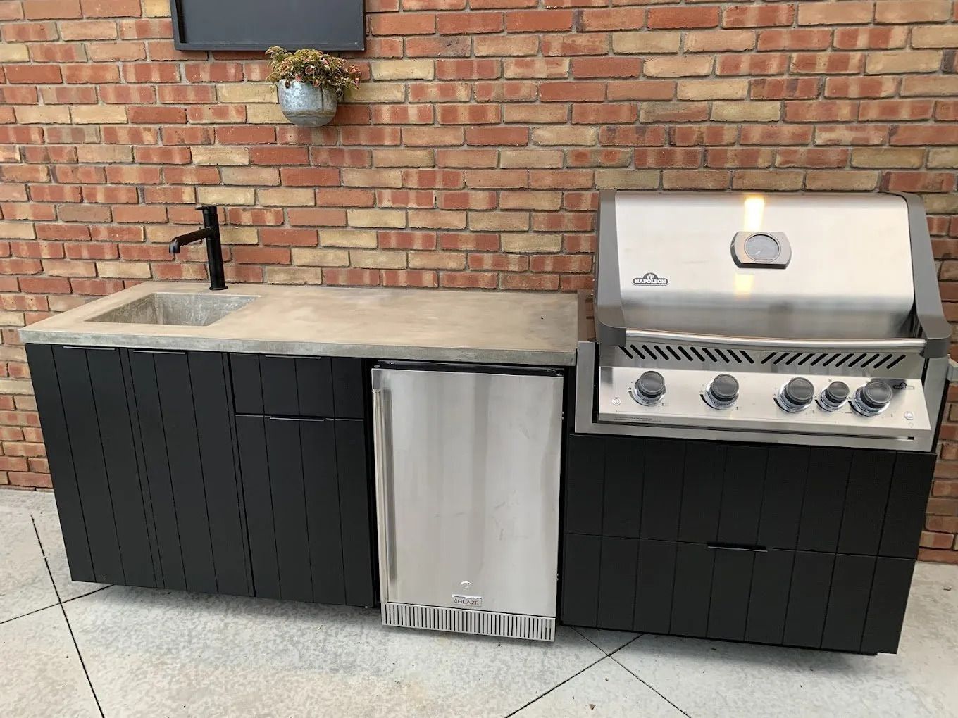 Outdoor kitchen with black cabinets, concrete countertop, stainless steel grill, refrigerator, and sink against a brick wall.