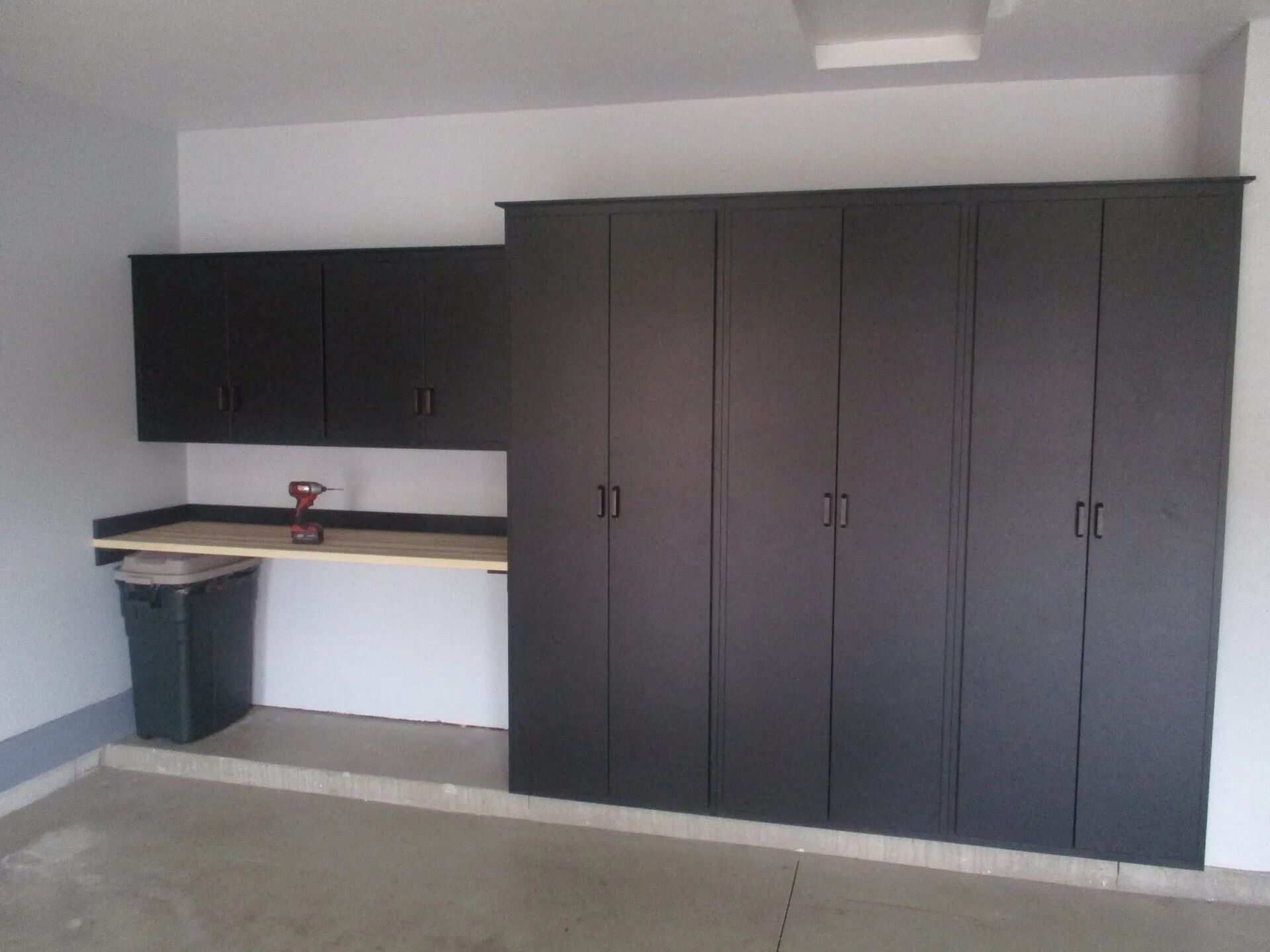 Dark cabinets and countertop in a garage setting, with a trash can and a power drill.