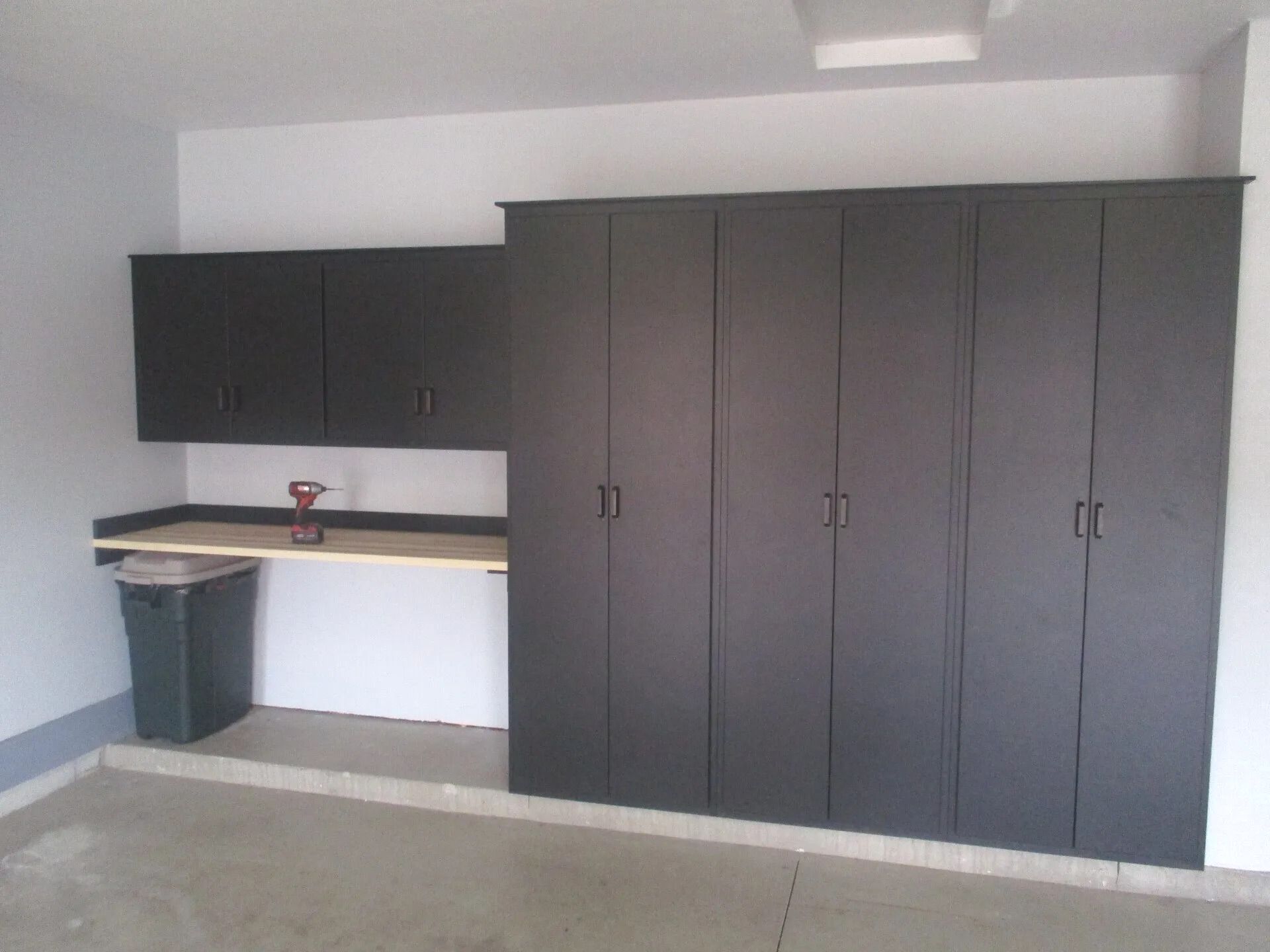Garage with dark cabinets, a workbench, and a trash can against a white wall and a concrete floor.