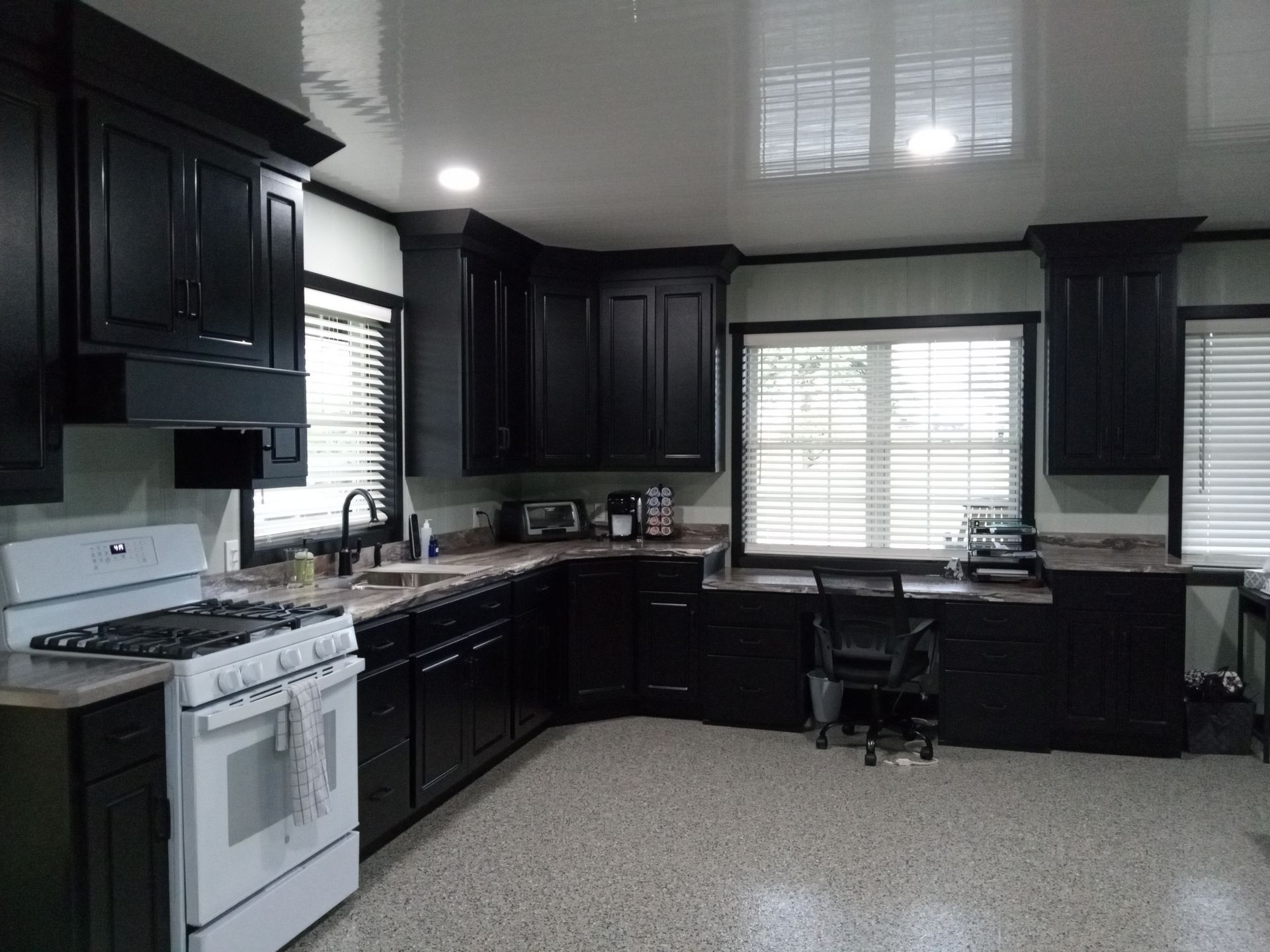 Black kitchen with white appliances, countertop, and cabinets. A window and desk are in the background.