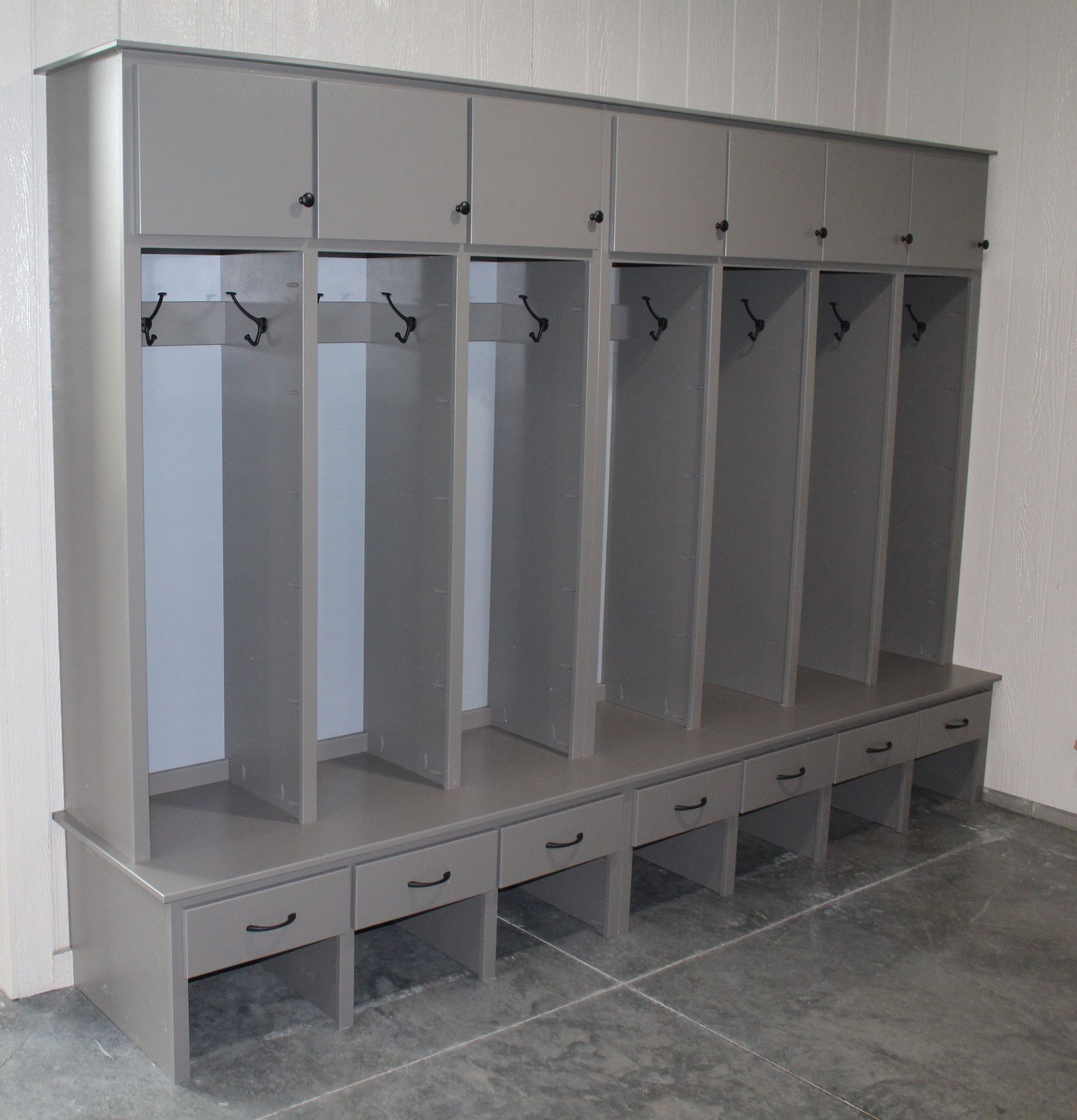 Gray lockers with hooks and overhead storage, sitting on a bench with drawers, against a light wall.