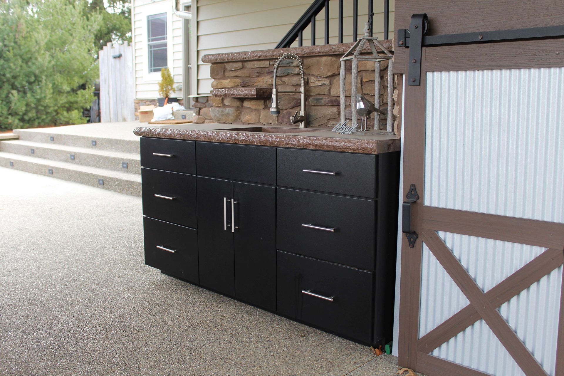 Outdoor black cabinet with sink, faucets, and stone backsplash, next to a barn door.