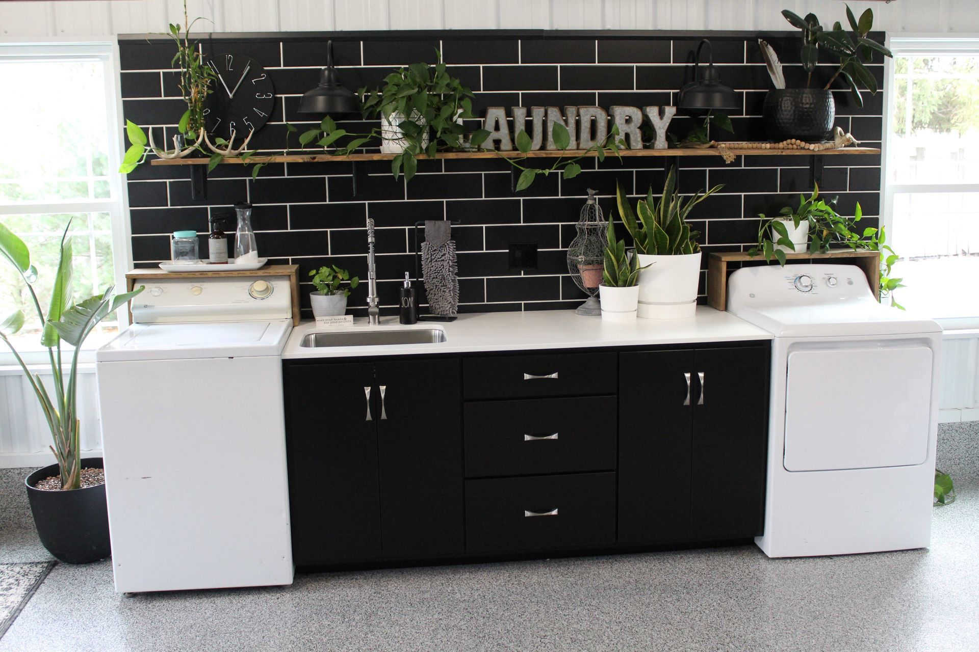 Laundry room with black brick wall, plants, white appliances, and a black cabinet.