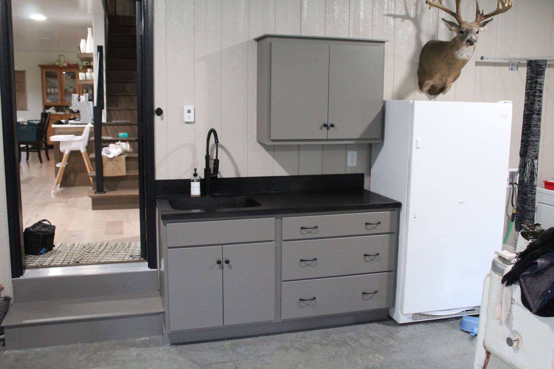 Gray cabinetry with a sink and a black countertop in a garage. A mounted deer head is visible.