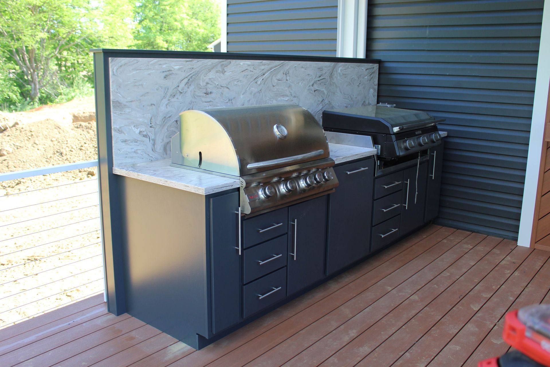 Outdoor kitchen with dark blue cabinets, gray countertop, two grills, and a stone backsplash on a wooden deck.