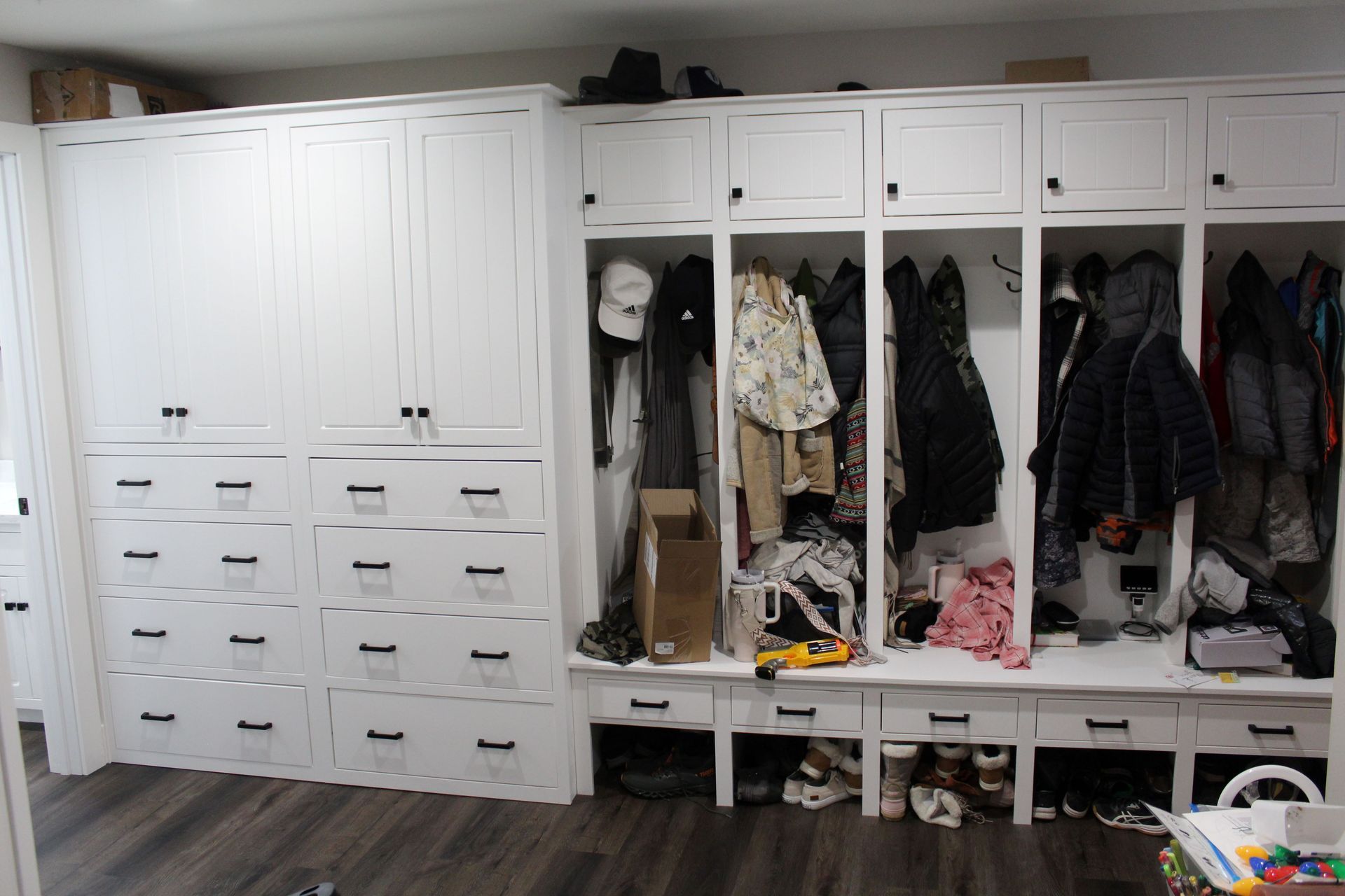 White built-in storage unit with drawers, cabinets, and open locker sections with coats, hats, and shoes.