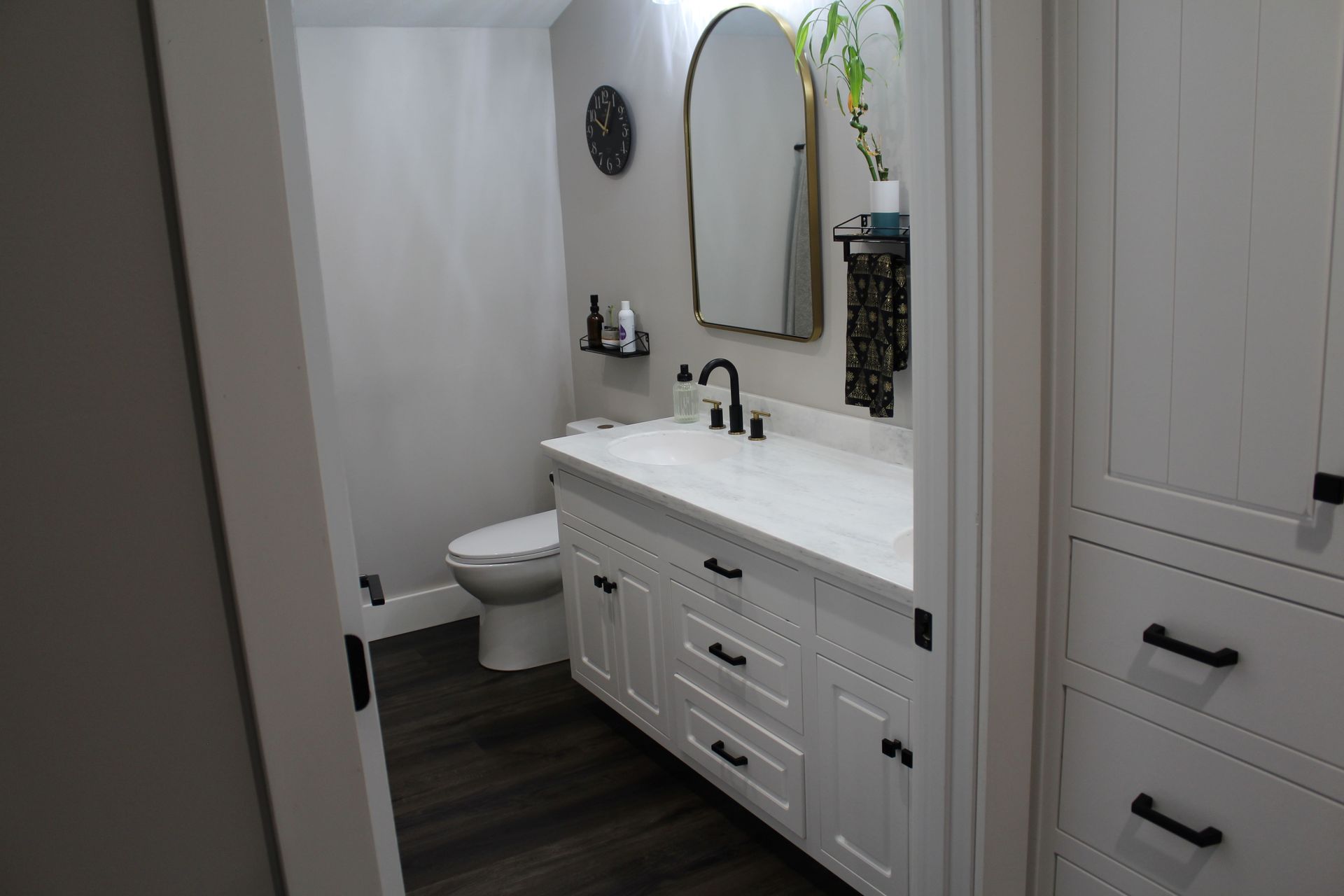 White bathroom with vanity, toilet, and mirror, featuring black hardware and dark flooring.