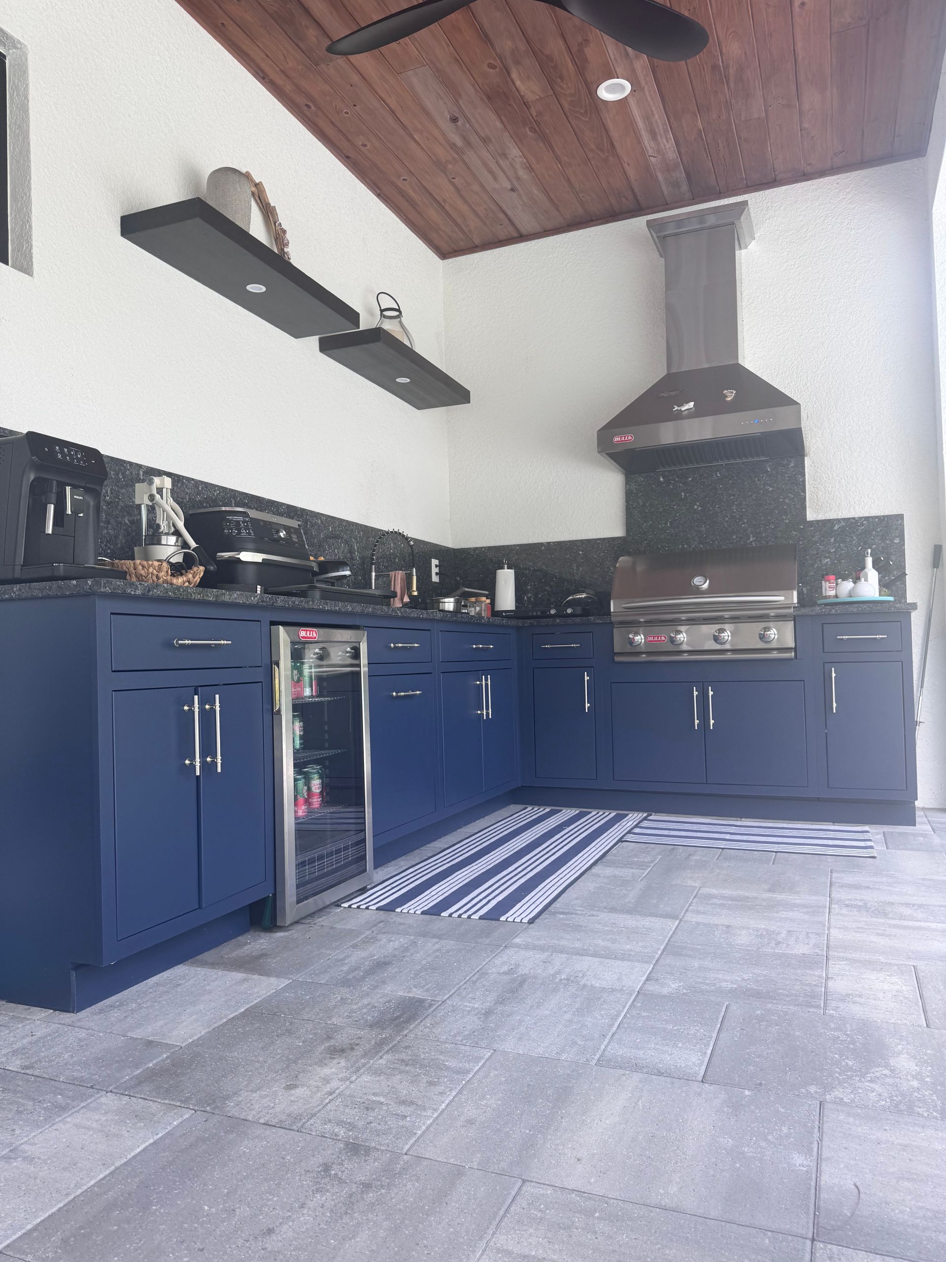 Outdoor kitchen with navy blue cabinets, stainless steel appliances, and a wood ceiling.
