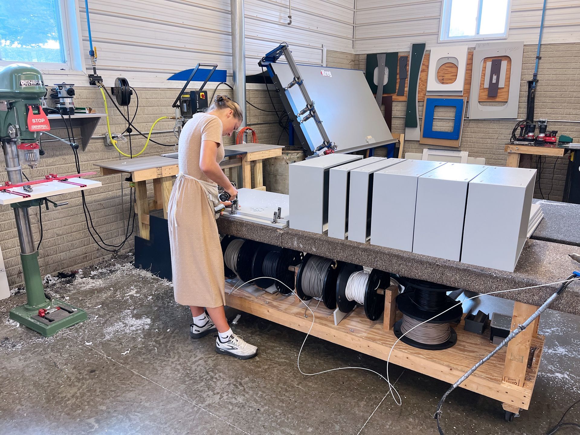 Woman working on a project in a workshop with tools, wires, and finished boxes on a worktable.
