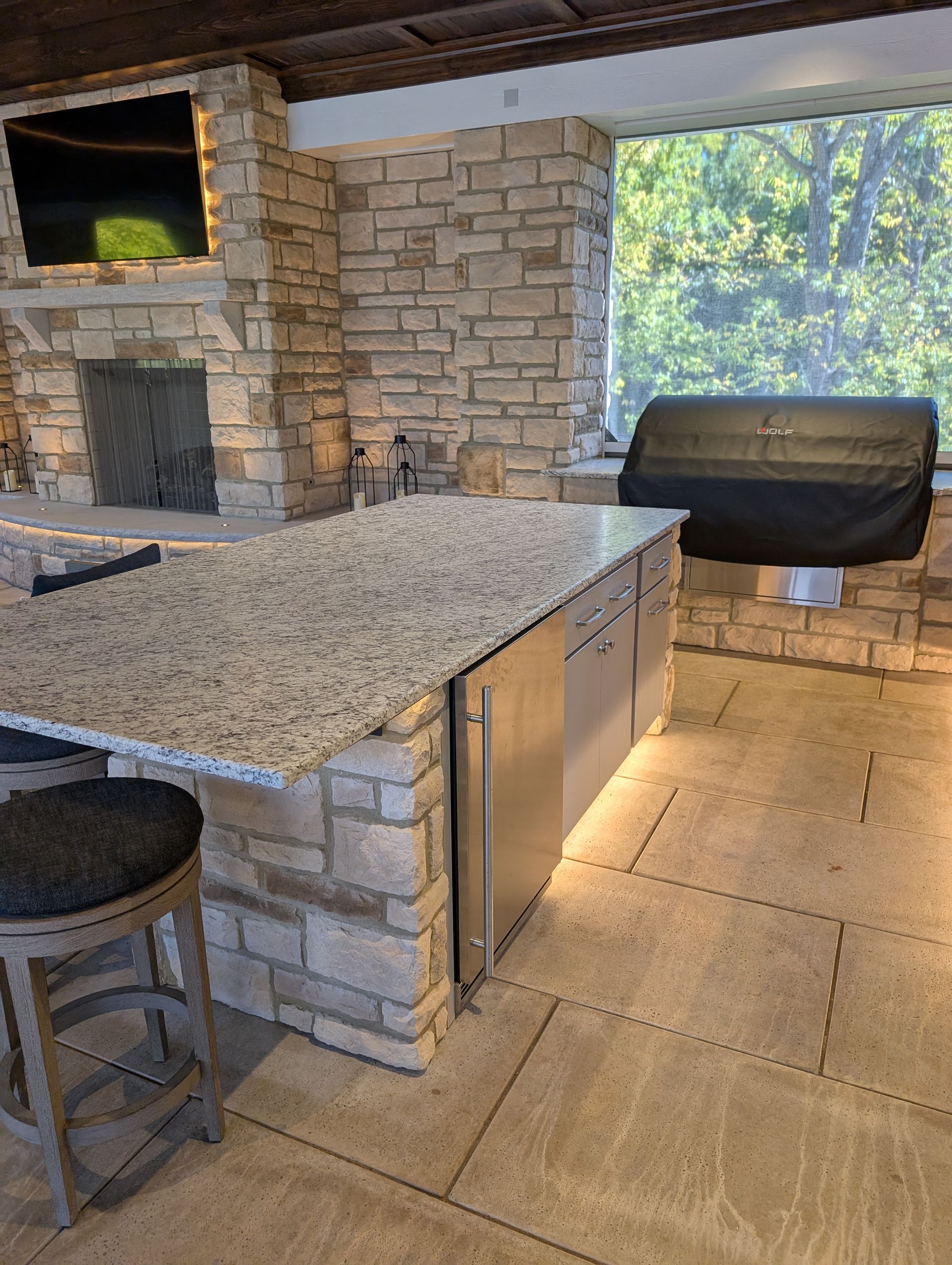 Outdoor kitchen with stone, granite island, appliances, and stool.